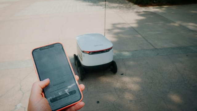 A person uses a smartphone to track an autonomous delivery robot in an outdoor setting.