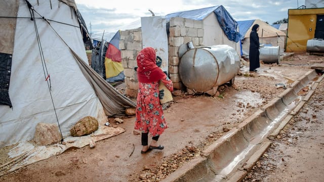Full body of poor women in hijabs and headscarves near weathered buildings and camps in village under cloudy sky