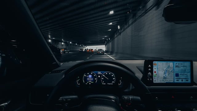 View from car dashboard driving through an illuminated city tunnel at night.