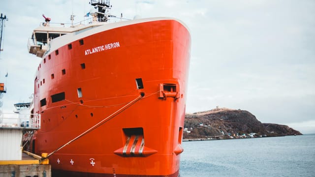 A striking red ship named Atlantic Heron is moored by a rocky shoreline on a clear day.