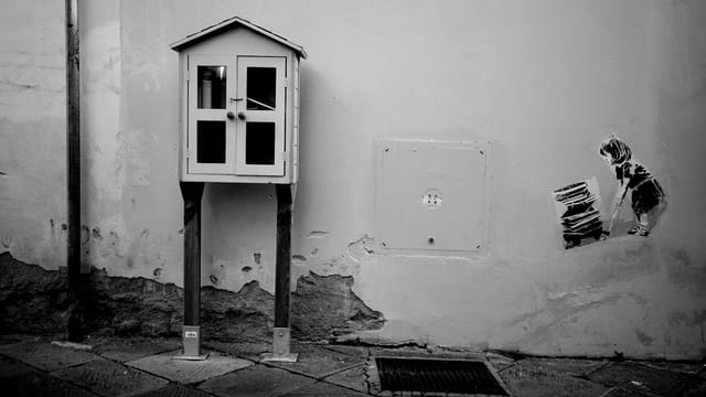 Monochrome image of street art showing a child next to a little free library on an urban wall.