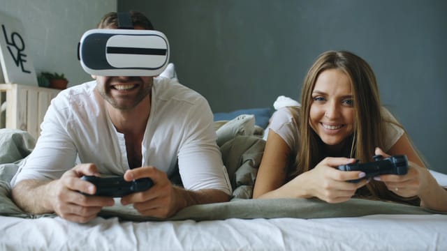Couple playing games in bed, one using VR headset. Relaxed indoor fun.
