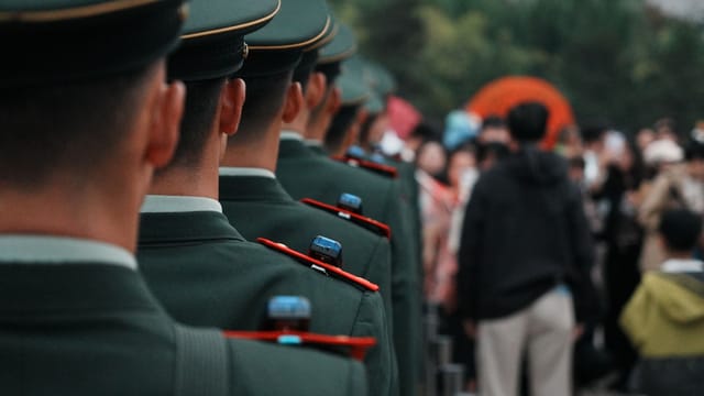 Military officers in green uniforms stand in line formation outdoors, facing a crowd.