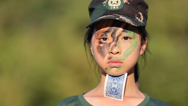 Young girl with face paint and cap, holding card, standing outdoors.