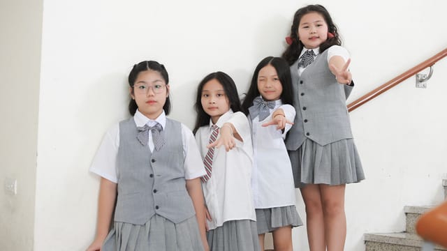 Four schoolgirls in grey uniforms stand on a staircase, smiling and posing.