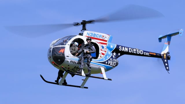 A police helicopter with armed personnel flying over San Diego on a clear day.
