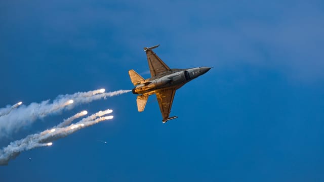 An F-16 fighter jet releases flares while performing an aerobatic maneuver against a clear blue sky at dusk.