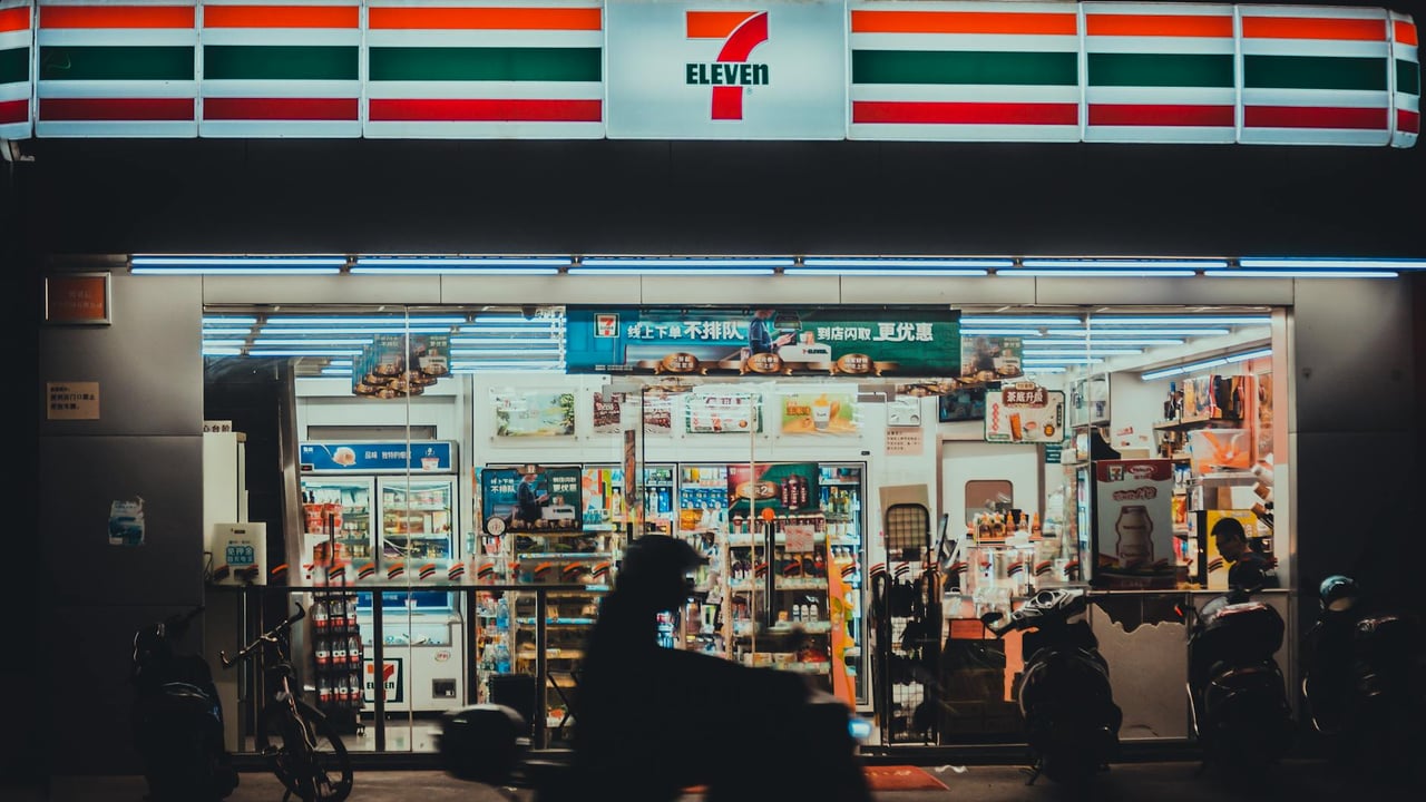 A vibrant 7-Eleven store front lit up during nighttime with a silhouette of a motorbike rider passing by.