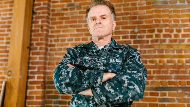 Proud US Navy veteran standing confidently in military uniform with brick background.