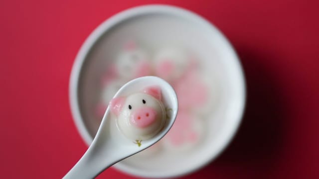 Adorable pig-shaped tang yuan in a ceramic spoon on a vibrant red background, perfect for festive occasions.