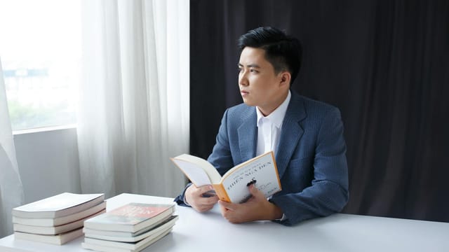 A young man in a suit thoughtfully reading a book at a table by the window.