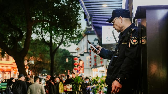 A street guard stands focused on his phone amid the bustling night streets of Nanjing, China.