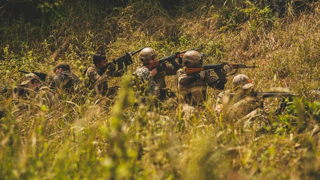 A group of soldiers in camouflage gear aim their weapons during a military exercise in a grassy field.