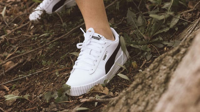 Close-up of white Puma shoes on person navigating through a forest trail with fallen leaves and branches.