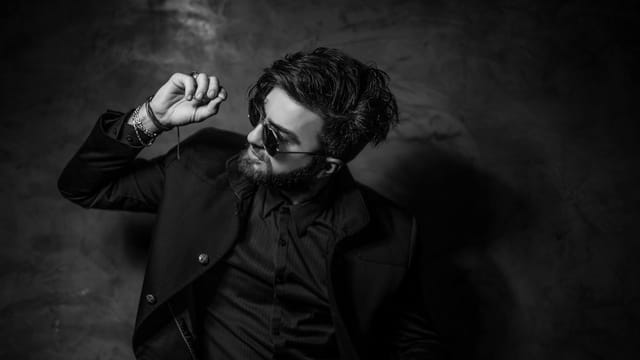 Black and white portrait of a fashionable man in sunglasses and suit posing confidently indoors.