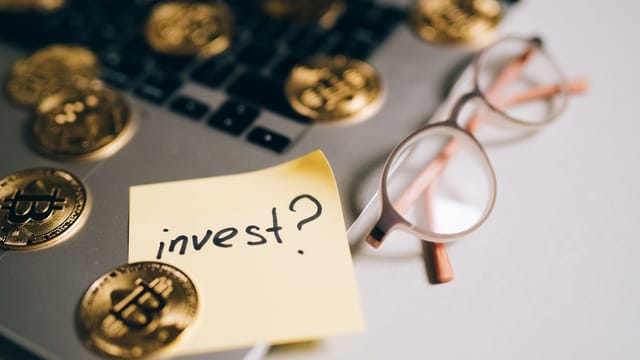 A close-up of bitcoin coins on a laptop with eyeglasses and a sticky note reading 'invest?'