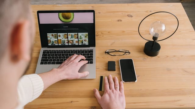 Man using laptop on wooden table with cellphone, bank card, and lamp nearby.