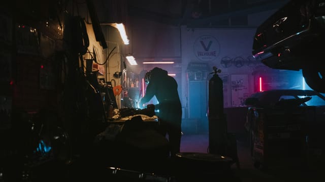 Silhouette of a mechanic working in a dimly lit garage, featuring neon lights and auto equipment.