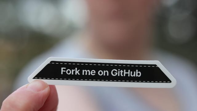 Close-up of a hand holding a 'Fork me on GitHub' sticker, blurred background.