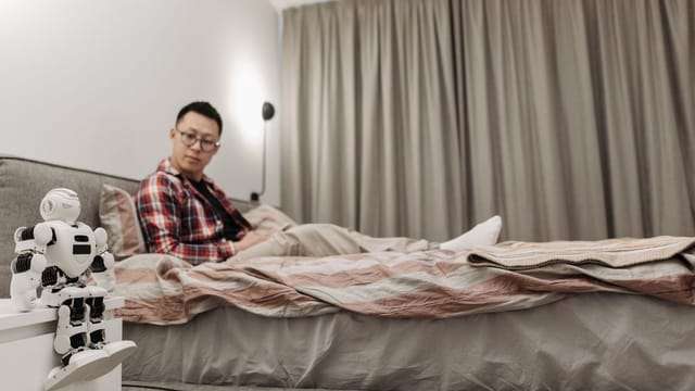 Asian man sitting in bedroom, observing futuristic robot on nightstand.