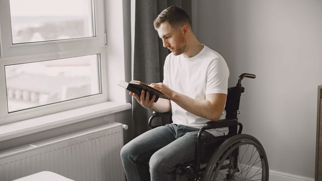 A man in a wheelchair reading a book in a well-lit room near a window.