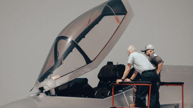 Two men inspecting military aircraft cockpit on a clear day.