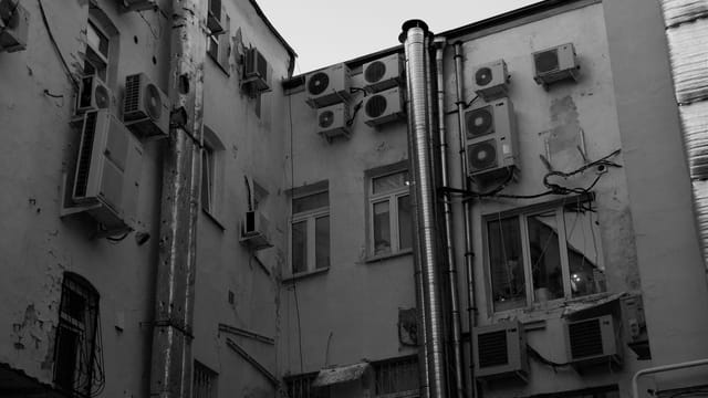 A black and white image of urban building exterior with multiple air conditioning units and pipes.