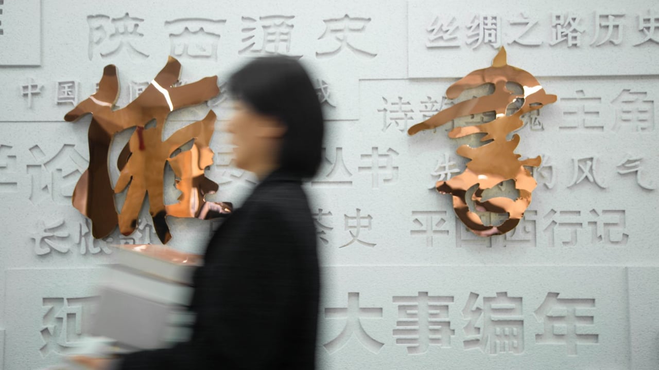 A blurred woman carries boxes past a wall with metallic Chinese characters, conveying motion and urban life.