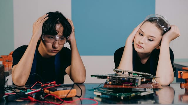 Two technicians in safety goggles work on circuit boards, displaying stress and focus in a tech lab.
