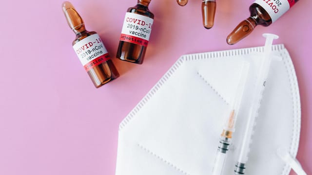 Close-up of COVID-19 vaccine vials, syringe, and mask on a pink surface.