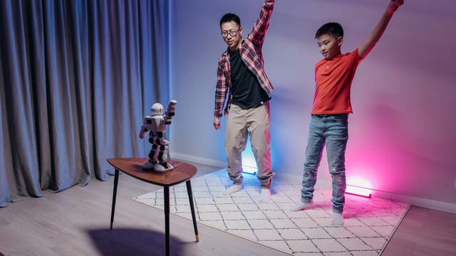 Father and son enjoy dancing with a humanoid robot in a colorful home setting.