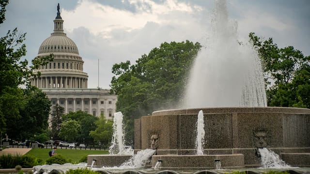 A scenic view of the US Capitol building with a fountain in the forefront, surrounded by lush greenery.