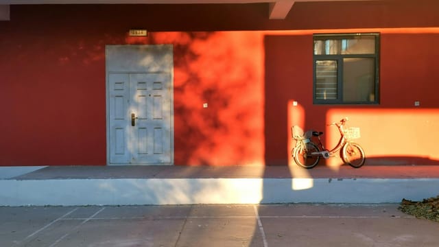 Bicycle parked against a vibrant red wall, casting shadows in warm sunlight, outdoors.