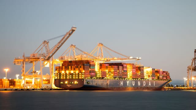 Container ship docked at port under cranes during twilight, illuminated against the evening sky.