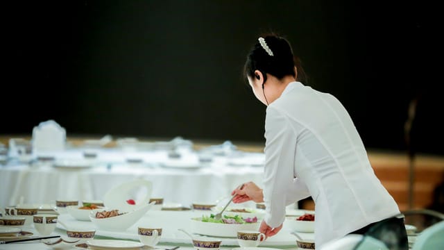 A waitress is seen preparing a table for a formal dinner setting indoors.