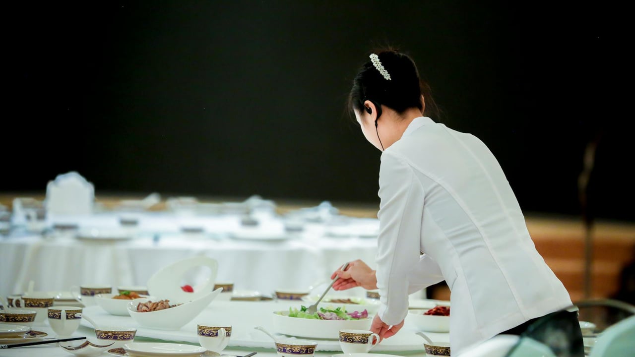 A waitress is seen preparing a table for a formal dinner setting indoors.