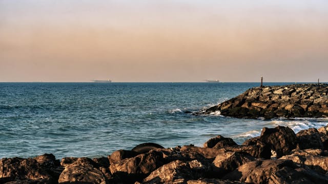 View of rocky shoreline with cargo ships in the distance along Sharjah coast, UAE.