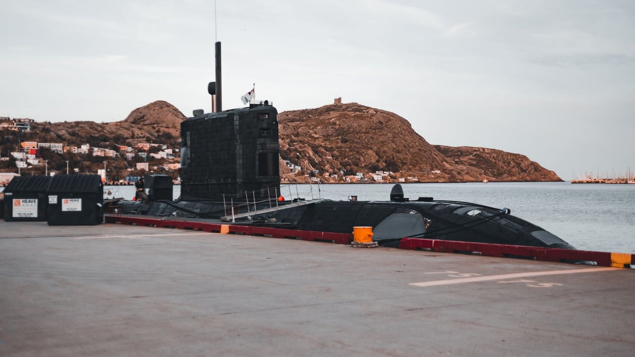 A submarine docked at an urban waterfront with scenic hills in the background.