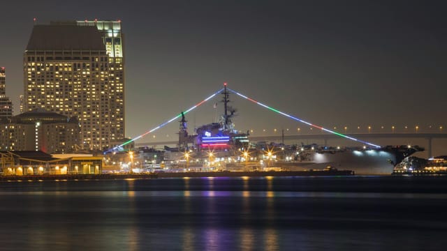 Night view of USS Midway Museum with city skyline, colorful lights in San Diego.
