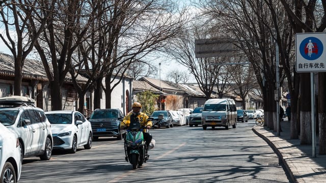 A motorcyclist rides through a bustling urban street lined with cars and bare trees.