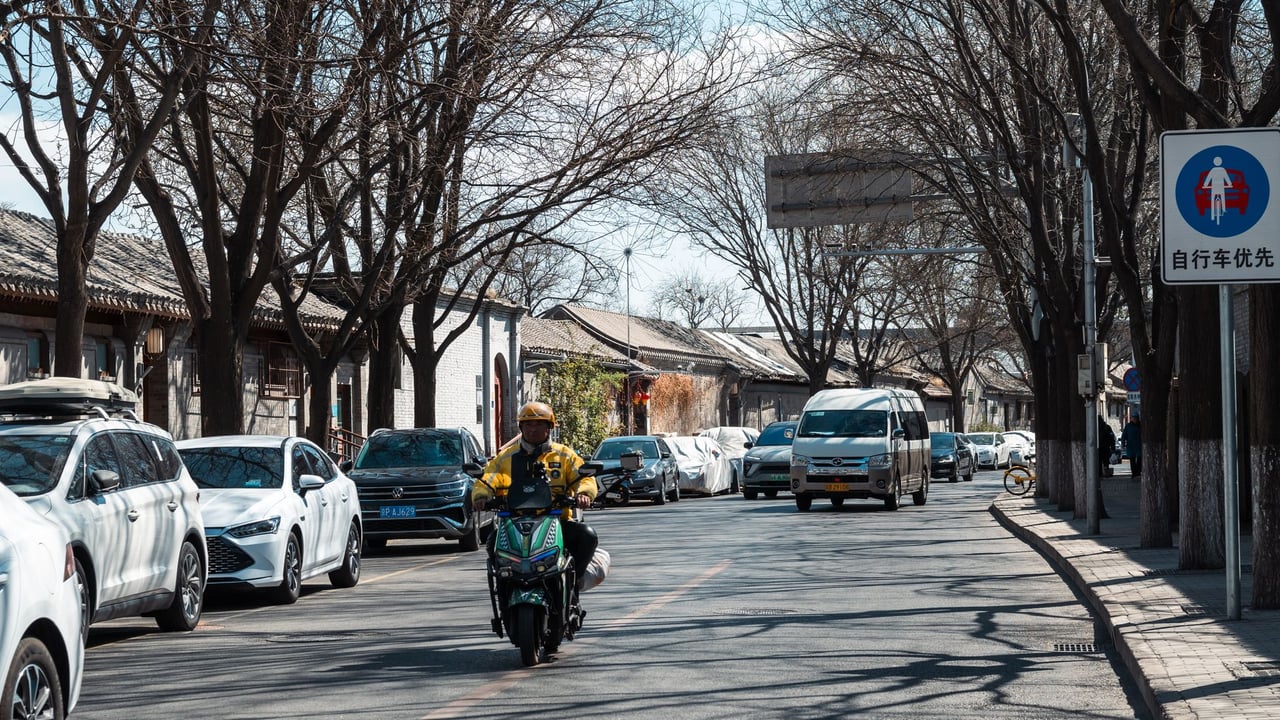 A motorcyclist rides through a bustling urban street lined with cars and bare trees.
