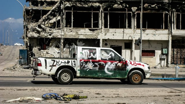 A damaged vehicle in front of a war-torn building in Sirte, Libya, depicting the impact of conflict.
