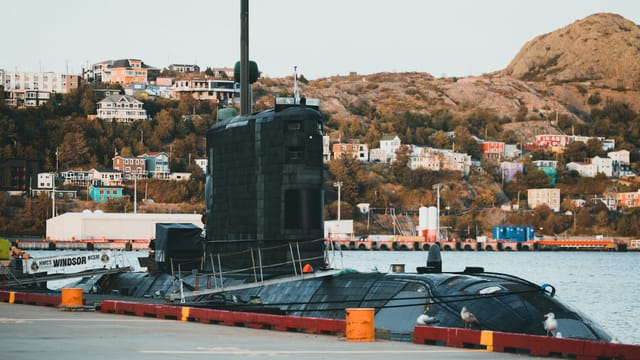 Submarine docked at a colorful hillside harbor with urban scenery.