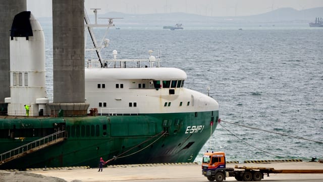 A green cargo ship docked at a busy industrial port with a visible truck on the dock.