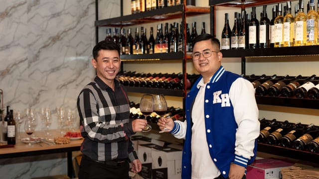 Two Asian men toast with wine glasses in a cozy wine cellar setting, surrounded by bottles.