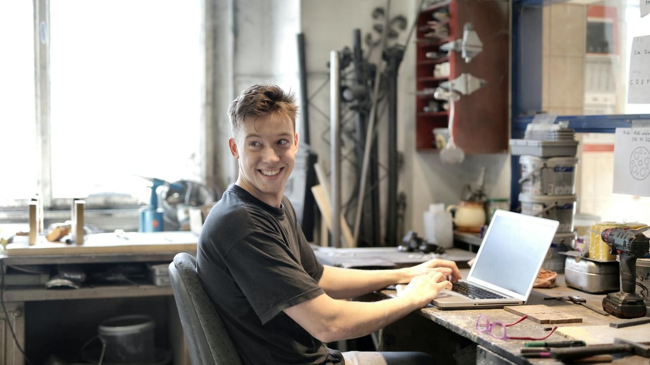 Young man smiling while using laptop in a creative workspace with tools around.
