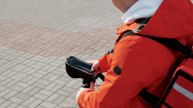 A food delivery courier in a bright red jacket checks his smartphone while riding a bicycle on a city street.