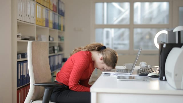Tired woman in red sweater naps on office desk beside laptop, overwhelmed by remote work.
