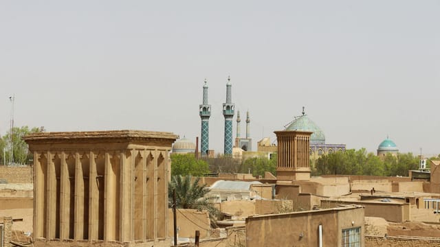View of Yazd's historic architecture with windcatchers and blue mosque domes under a clear sky.