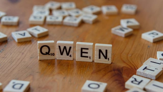 Wooden Scrabble tiles form the word 'QWEN' on a wooden surface, with scattered tiles in the background.
